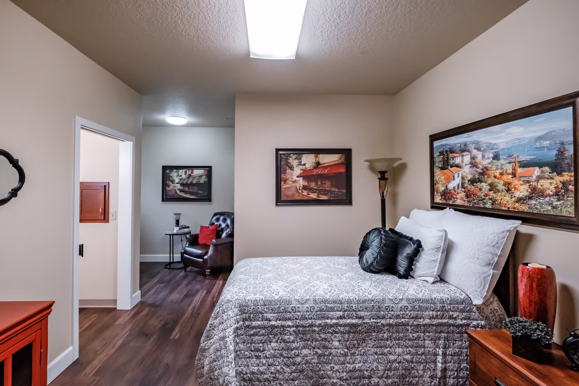 A cozy bedroom in a senior living facility with a bed covered in a patterned gray and white bedspread, adorned with white and black pillows. The room features wooden flooring, a red cabinet, a wooden nightstand with a red vase and a small plant, and a tall floor lamp. Two framed paintings hang on the walls, one above the bed depicting a scenic landscape and another on the adjacent wall showing a café scene. In the background, there is a small sitting area with a dark leather armchair and a side table with a lamp.
