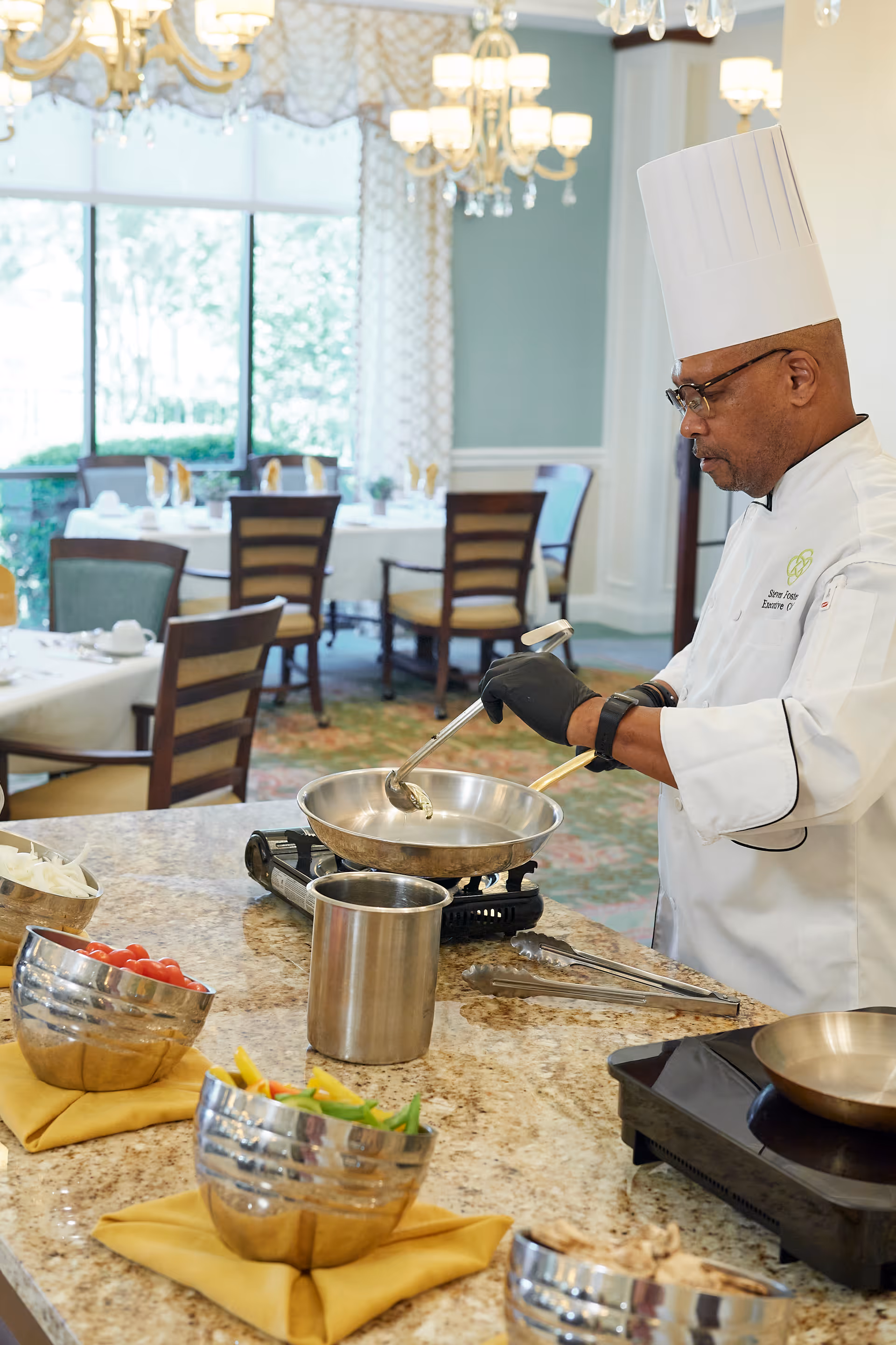 A chef wearing a white uniform and tall white hat is cooking with a ladle in a pan on a portable stove in a dining area. The countertop in front of him has bowls of fresh vegetables and utensils. The background shows tables and chairs set for dining with large windows letting in natural light.