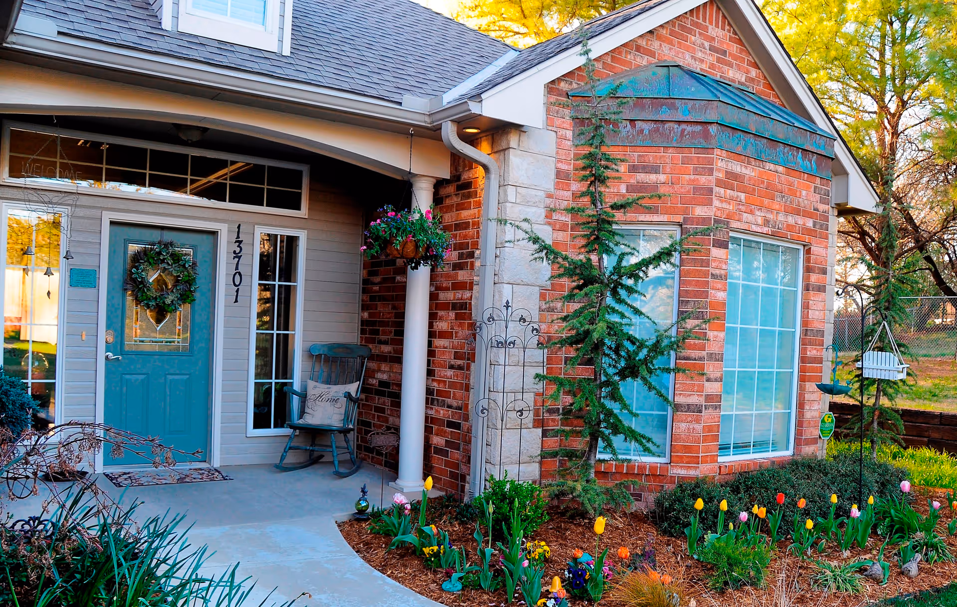 Front entrance of a brick house with a teal door, porch chair, hanging planter, and a colorful flower garden.