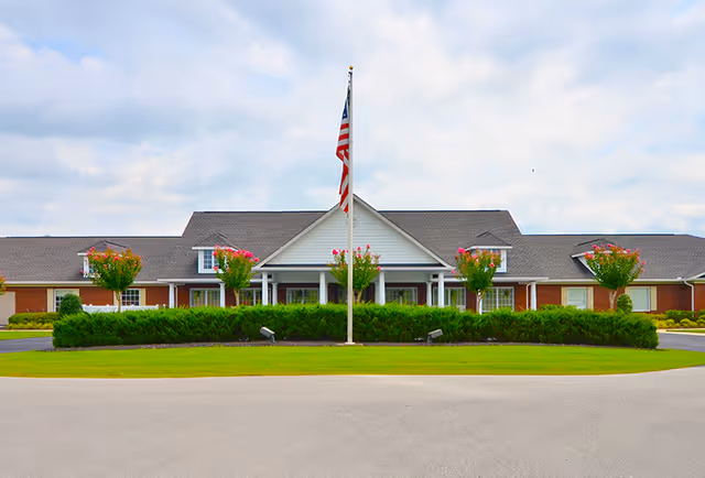 Front exterior of a single-story senior living building with a centered flagpole and landscaped circular driveway.