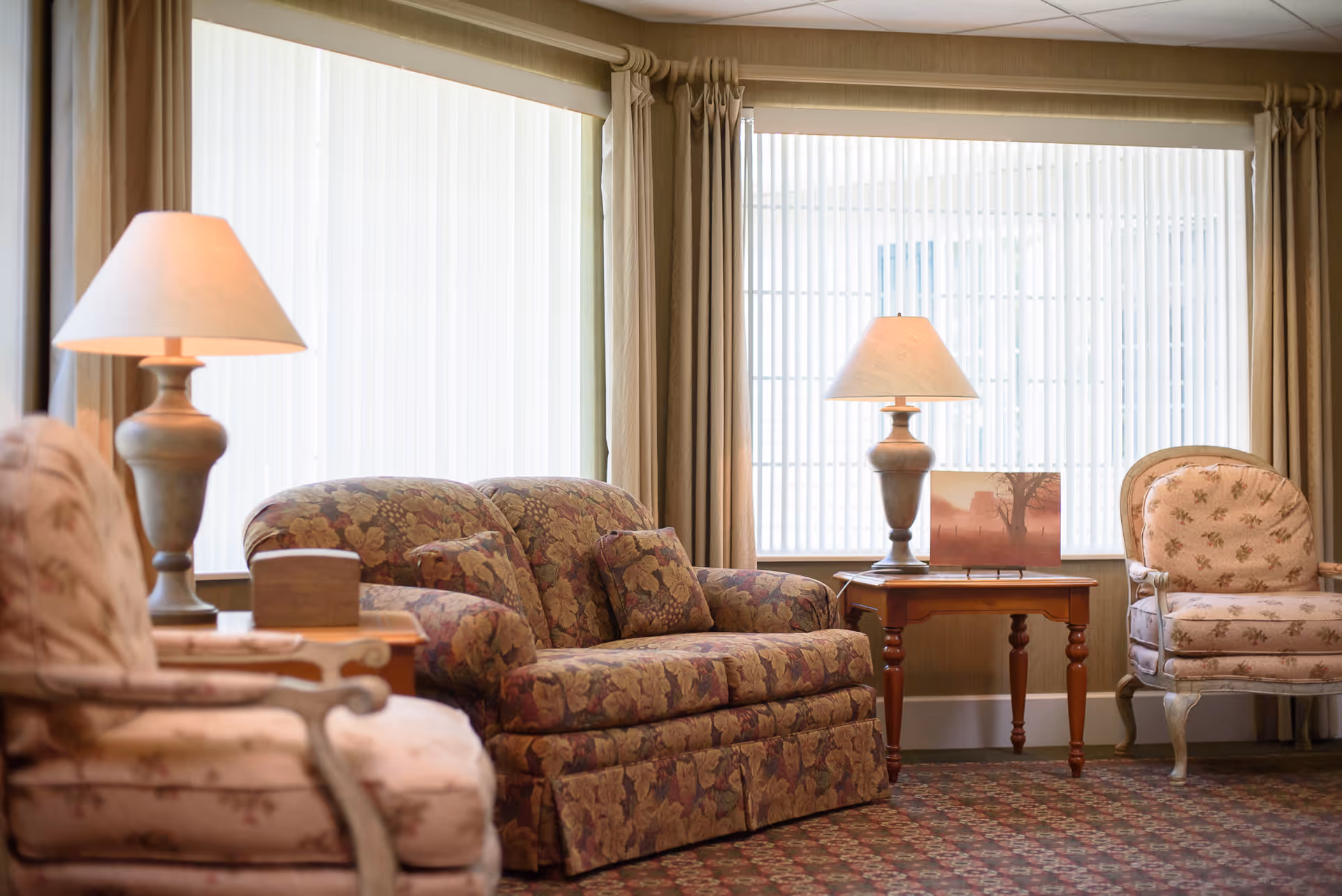 A cozy living room area with floral upholstered furniture including a loveseat and two armchairs. There are two table lamps on wooden side tables, and large windows with vertical blinds and beige curtains letting in natural light. A framed picture is placed on one of the side tables.
