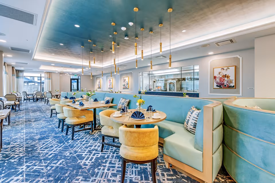 A bright and modern dining room with round wooden tables set with blue napkins, white cups, and glassware. The seating includes light yellow cushioned chairs and curved turquoise banquettes with patterned pillows. The ceiling features a dark blue accent with multiple gold pendant lights hanging down. The room has large windows with curtains, patterned blue carpet, and framed floral artwork on the walls. A glass window reveals a kitchen area in the background.