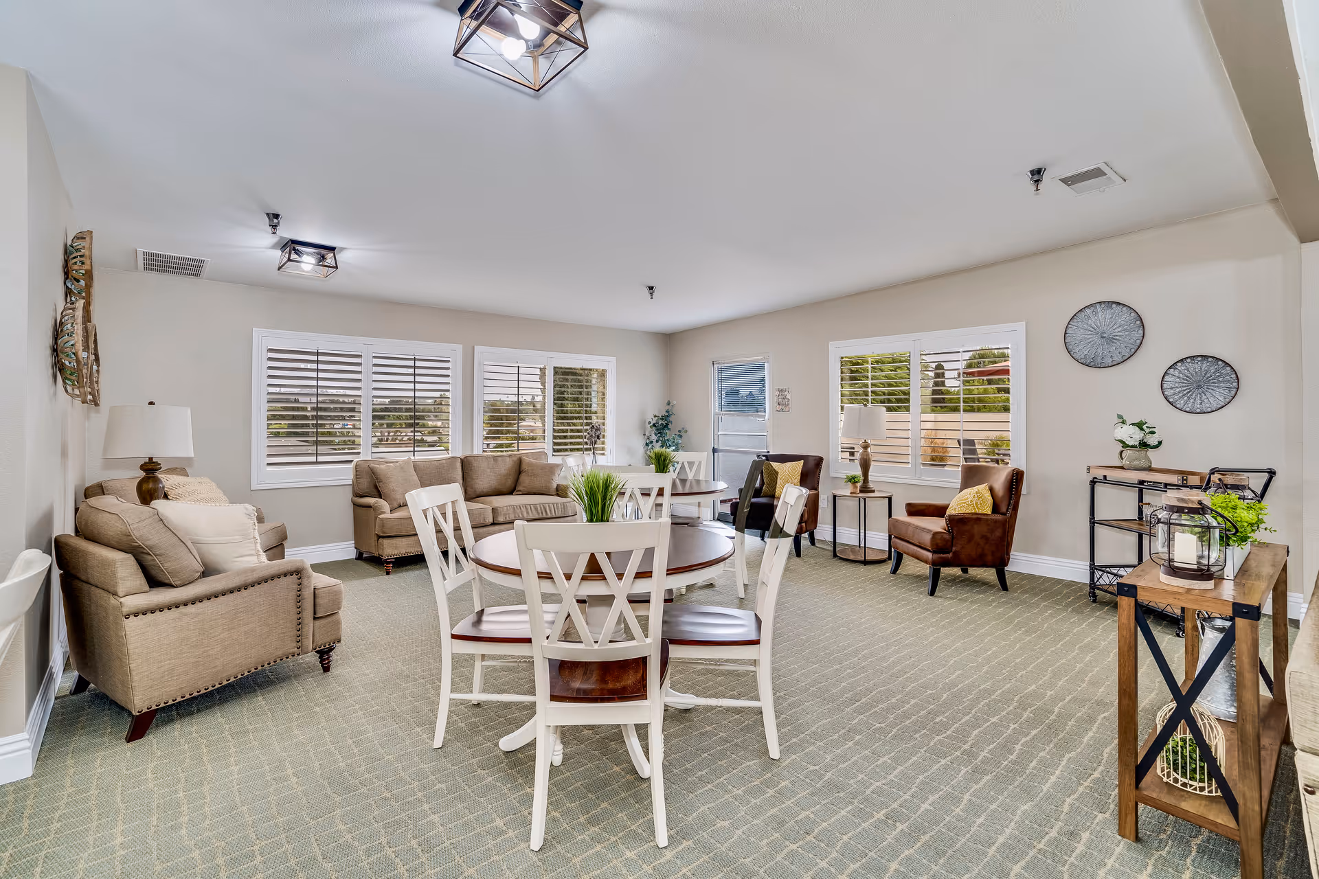 A bright and spacious living room area in an assisted living facility featuring beige walls and carpeted floor. The room contains a round wooden table with four white chairs in the center, a beige sofa and armchair on the left, and two brown leather armchairs near the windows on the right. The windows have white shutters, and there are decorative items such as plants, wall art, and lamps throughout the room.