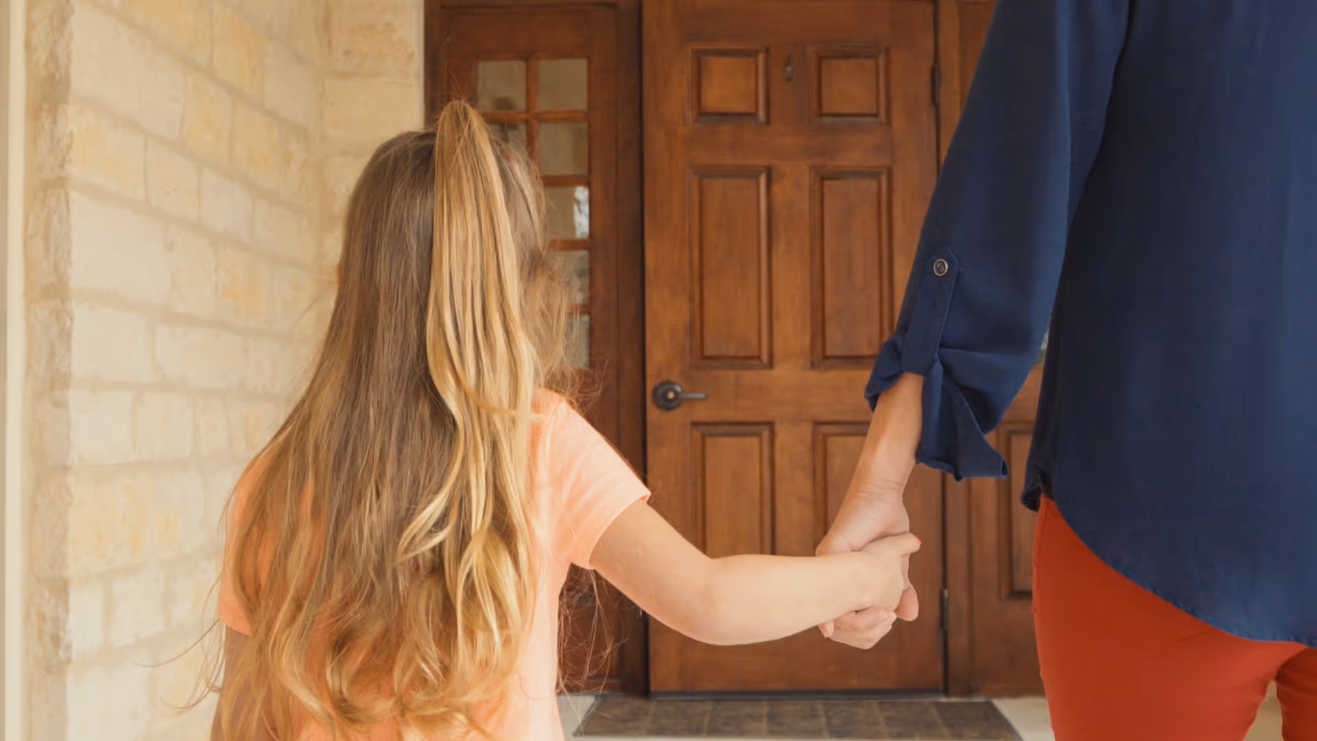 A young girl with long blonde hair holding hands with an adult, standing in front of a wooden door with glass panels on the side, inside a stone-walled entryway.
