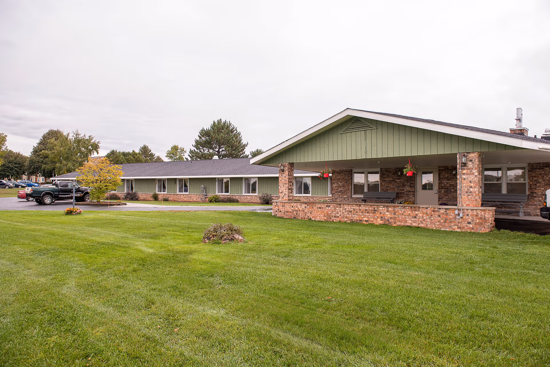 Exterior view of a single-story senior living facility building with green siding and brick accents. The building has a covered entrance with hanging flower pots and benches. There is a well-maintained green lawn in the foreground and several parked vehicles to the left side of the building under a cloudy sky.