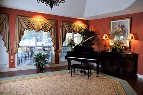 A cozy living room area with a grand piano and piano bench on a large patterned rug. The walls are painted a warm reddish color and decorated with a framed floral painting and two wall lamps with shades. There are three large windows with striped valances and white blinds, and a potted plant near the windows. A dark wooden sideboard with flower arrangements is placed against the wall.