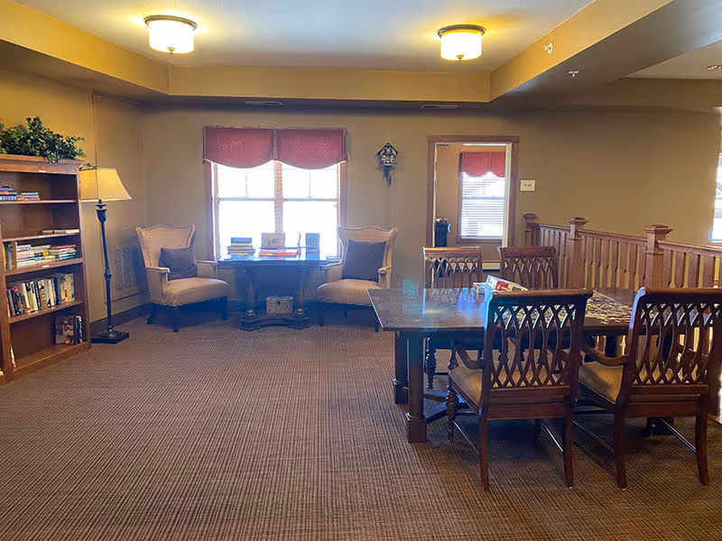 A cozy senior living common area with a wooden bookshelf filled with books on the left, two cushioned armchairs with pillows facing a small table in front of a window with red valances. To the right, there is a wooden dining table with six chairs. The room has beige walls, carpeted floor, and warm ceiling lights.