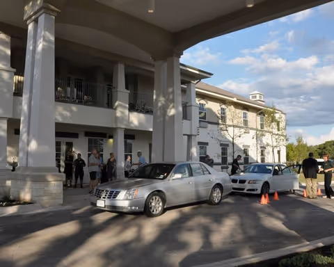 Entrance area of a senior living facility with a covered driveway where several people are standing and conversing near parked cars under a large portico attached to a two-story building.