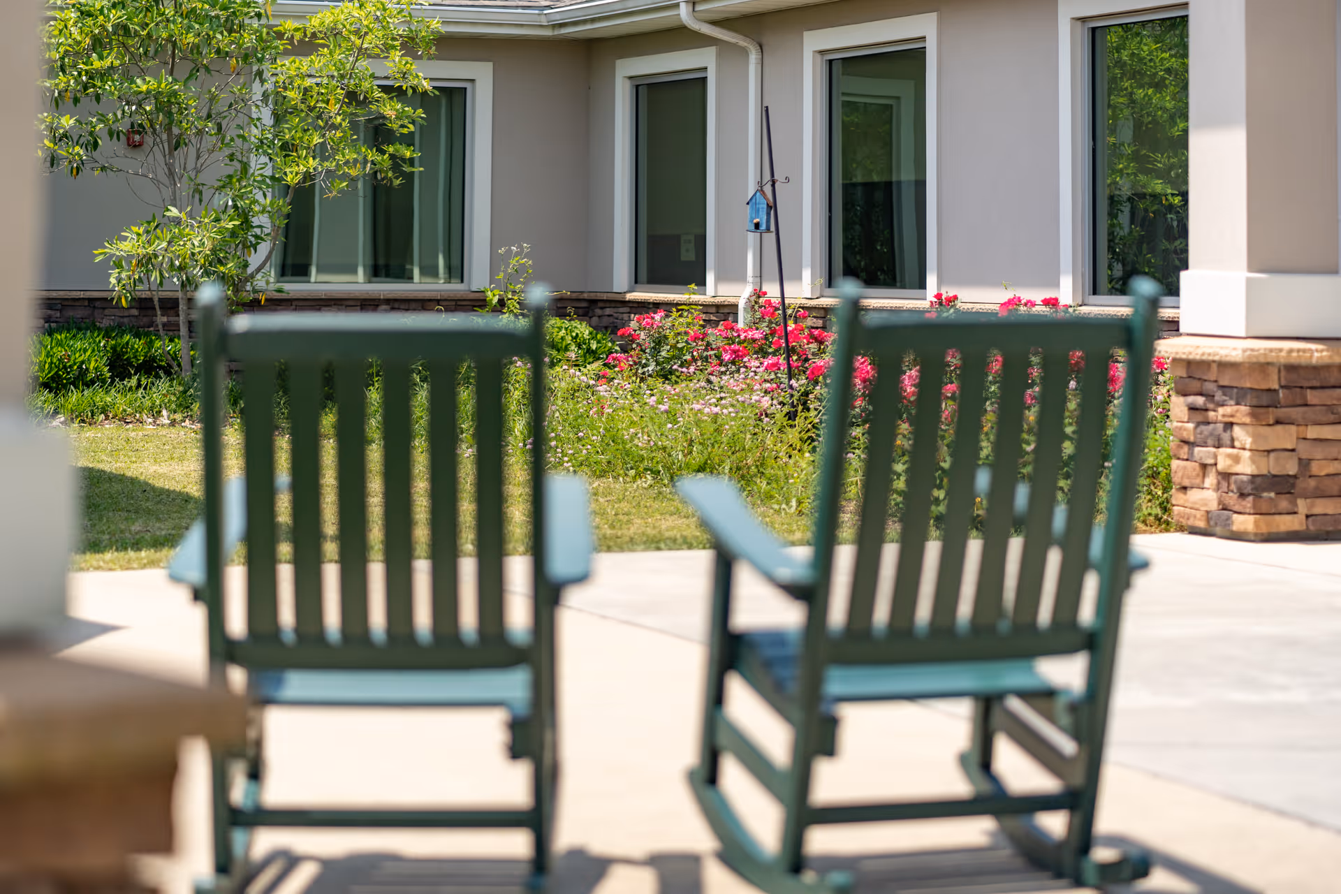 Two green rocking chairs on a sunny patio facing a landscaped garden and the exterior of a senior living building.