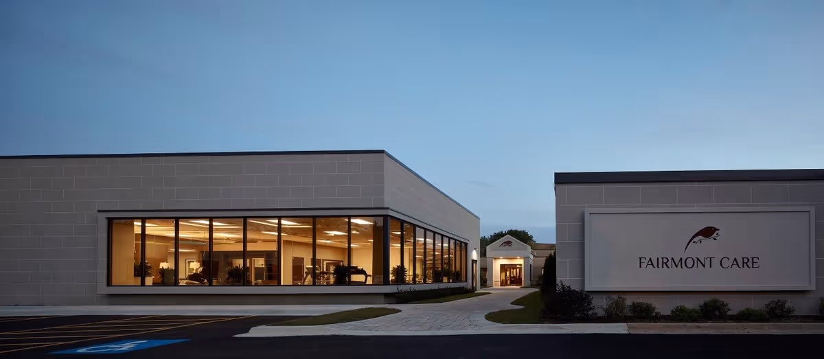 Exterior view of a modern single-story building at dusk with large windows showing a well-lit interior. A pathway leads to an entrance under a small portico. A sign on the right side of the image reads 'FAIRMONT CARE'.