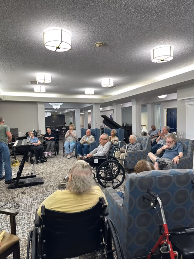 A group of elderly residents seated in a spacious, well-lit common area with carpeted floors and modern ceiling lights. Some residents are in wheelchairs, others in armchairs, attentively watching a man playing an electronic keyboard. The room features white columns and a grand piano in the background.