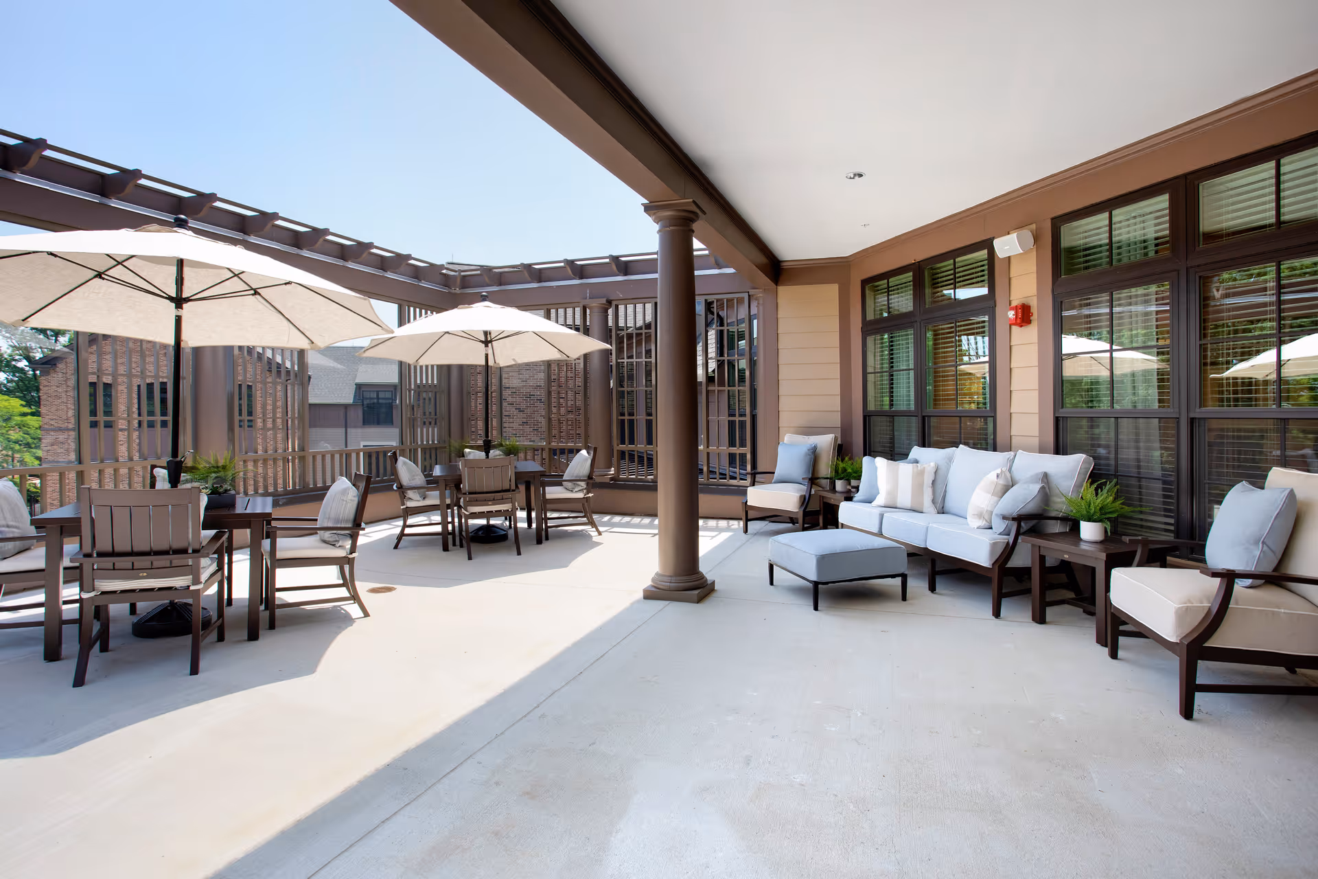 Covered outdoor patio with tables, umbrellas and cushioned lounge seating outside a senior living building.