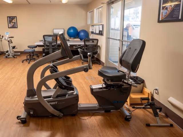 Indoor exercise room with a recumbent stationary bike in the foreground, two blue exercise balls on a desk in the background, office chairs, and a stationary bike near the wall. The room has wooden flooring and beige walls with framed posters.