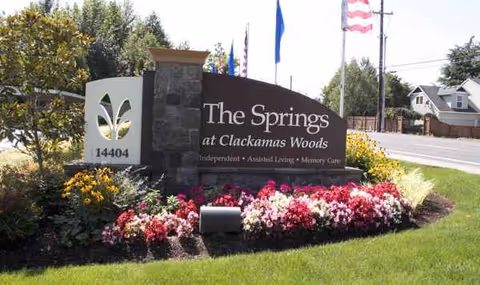 Entrance sign for The Springs at Clackamas Woods with flower beds and flags.