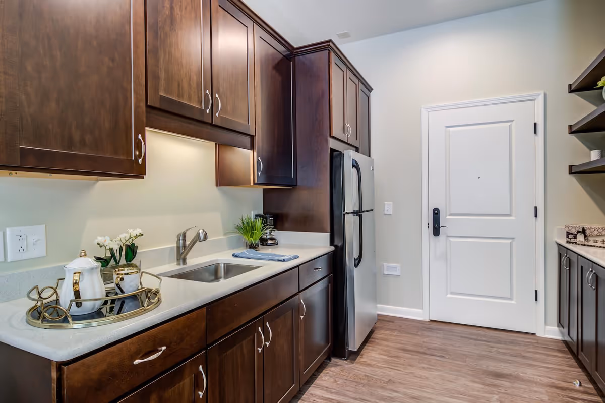 A modern kitchen with dark wooden cabinets, a stainless steel refrigerator, a sink with a faucet, and a white countertop. There is a decorative tray with a teapot and cups, a small plant, and a coffee maker on the counter. The floor is wooden, and there is a white door at the end of the kitchen.