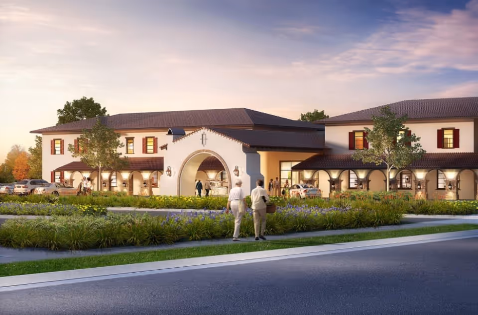 Exterior view of a two-story senior living facility with a Spanish-style design, featuring arched entryways, red shutters, and a tiled roof. There are landscaped gardens with green shrubs and purple flowers in front, a paved road, and several people walking near the entrance. Cars are parked along the side of the building under covered areas.
