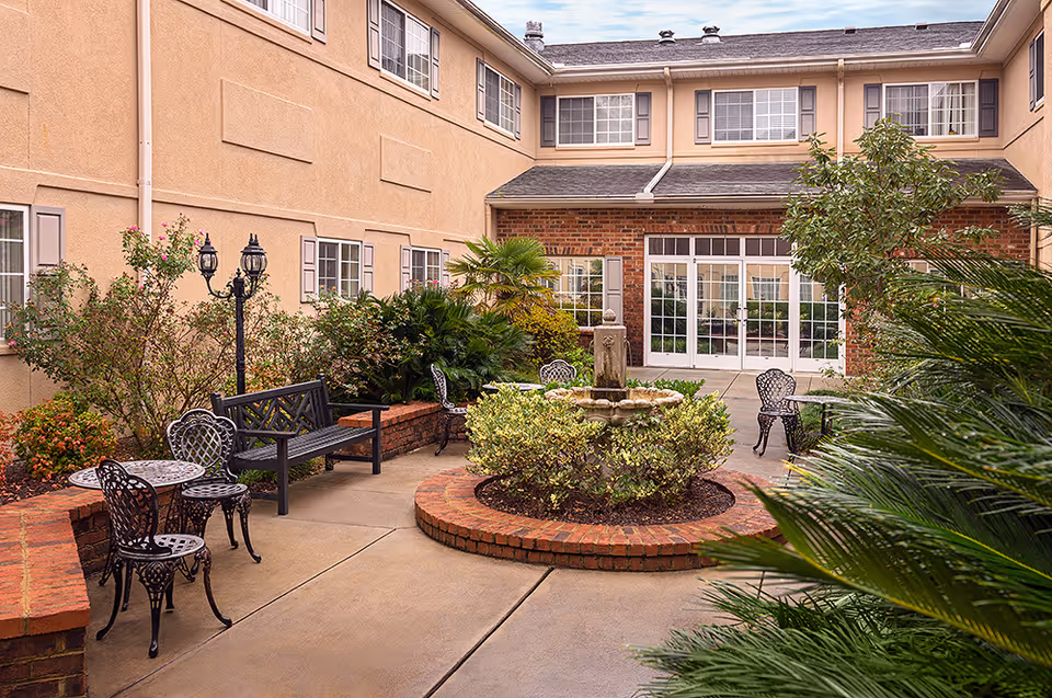 Enclosed courtyard with a central fountain, wrought-iron tables and chairs, benches and surrounding building facade with windows and glass doors.