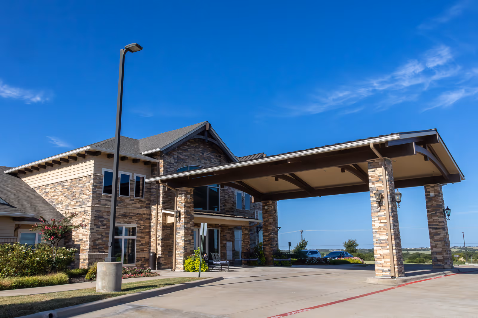 Front entrance of a stone-faced assisted living building with a covered porte-cochère under a clear blue sky.