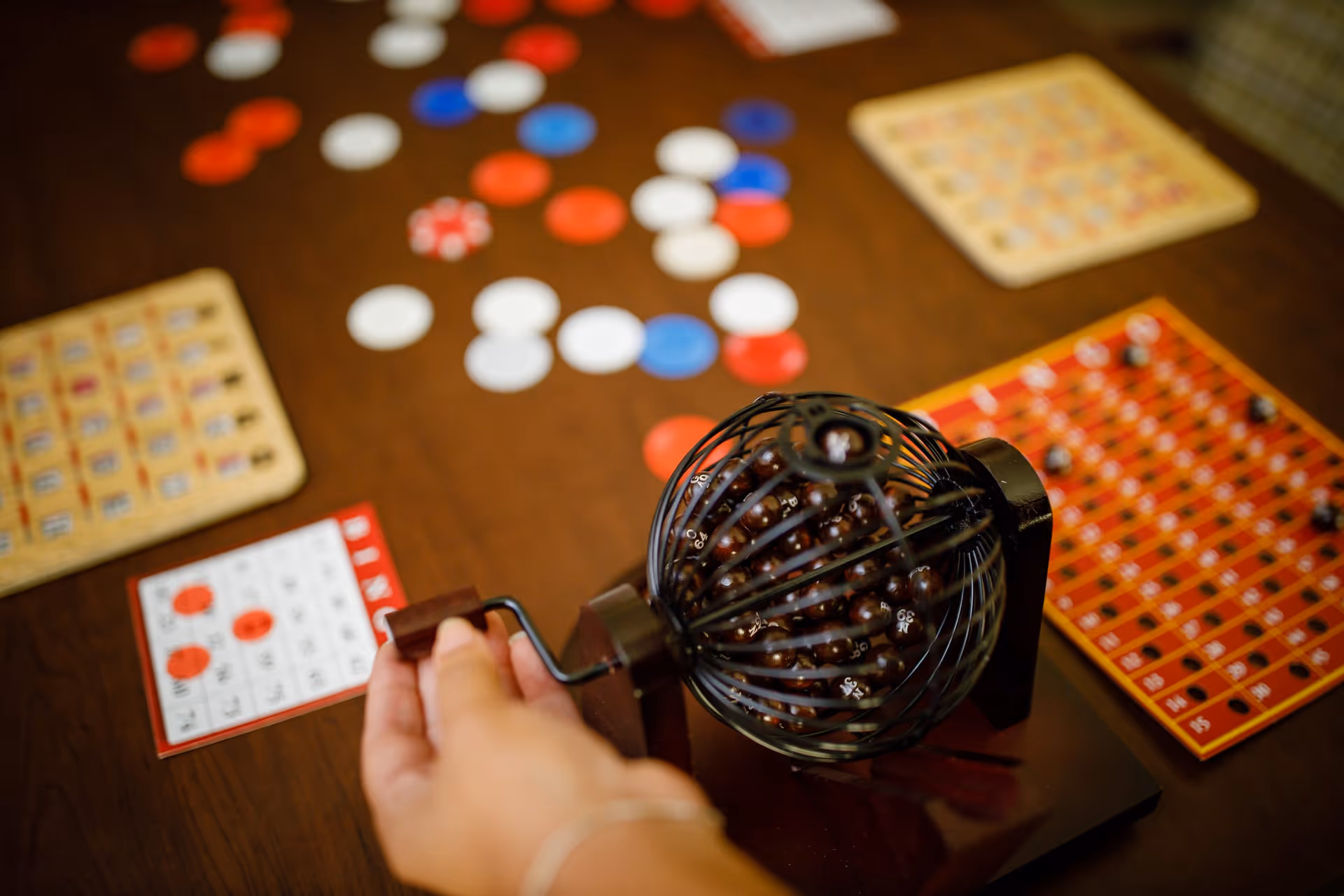 A close-up of a hand turning a bingo cage with numbered balls inside on a wooden table, surrounded by bingo cards and red, white, and blue bingo chips scattered around.