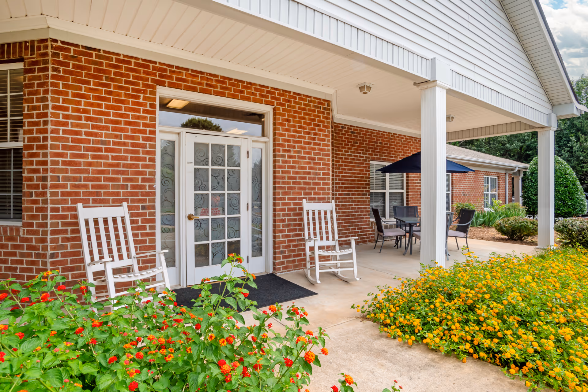 Covered brick entrance with glass double doors, white rocking chairs, a patio table with umbrella, and flowering shrubs.