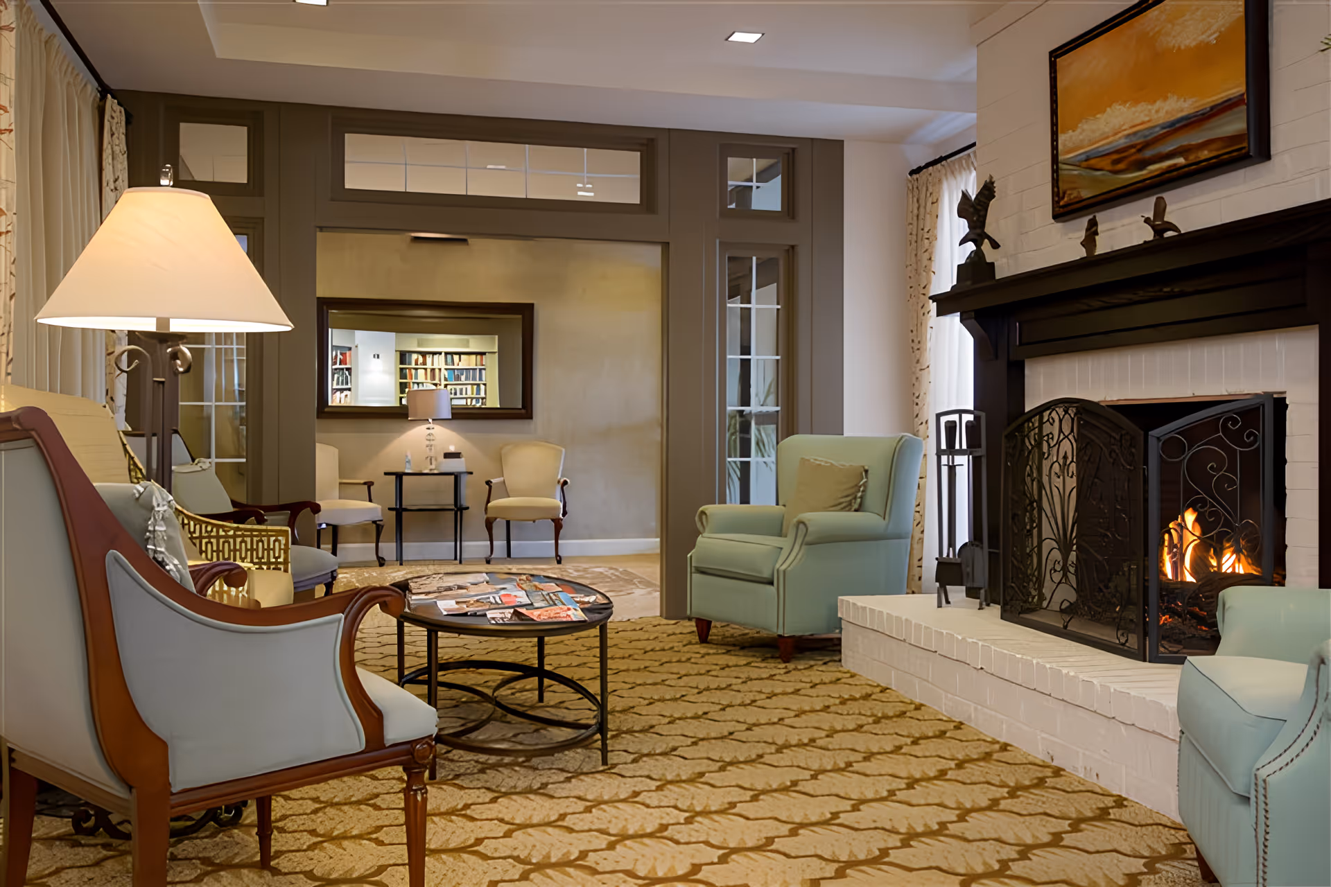Cozy furnished common living room with upholstered chairs, a coffee table, and a lit fireplace.