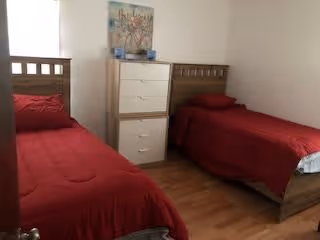 A small bedroom with two single beds, each with a red bedspread. Between the beds is a white chest of drawers with a decorative picture and some items on top. The room has wooden flooring and plain white walls.