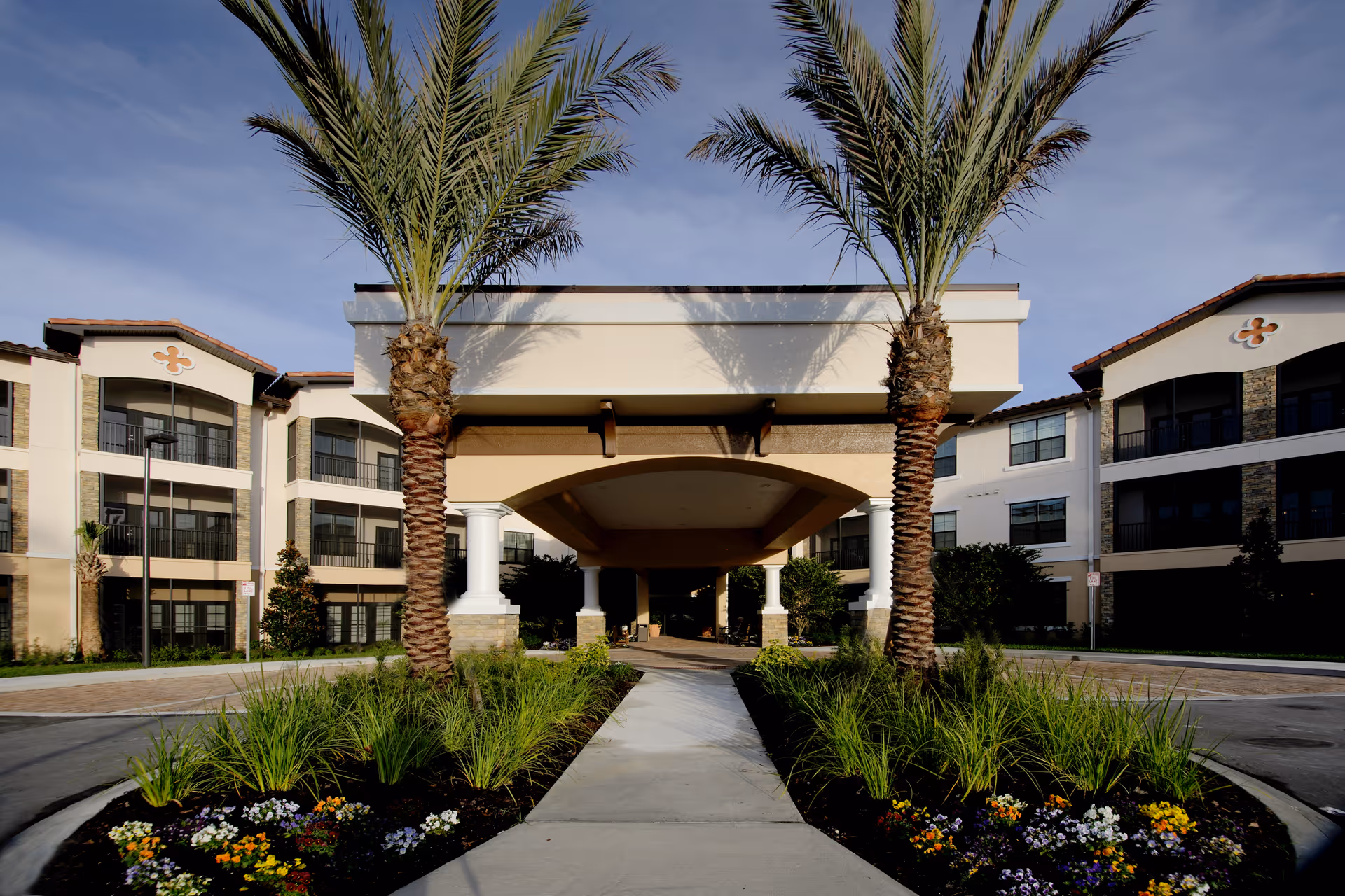 Front entrance of a senior living building with a covered porte-cochere, palm trees, and landscaped flower beds.