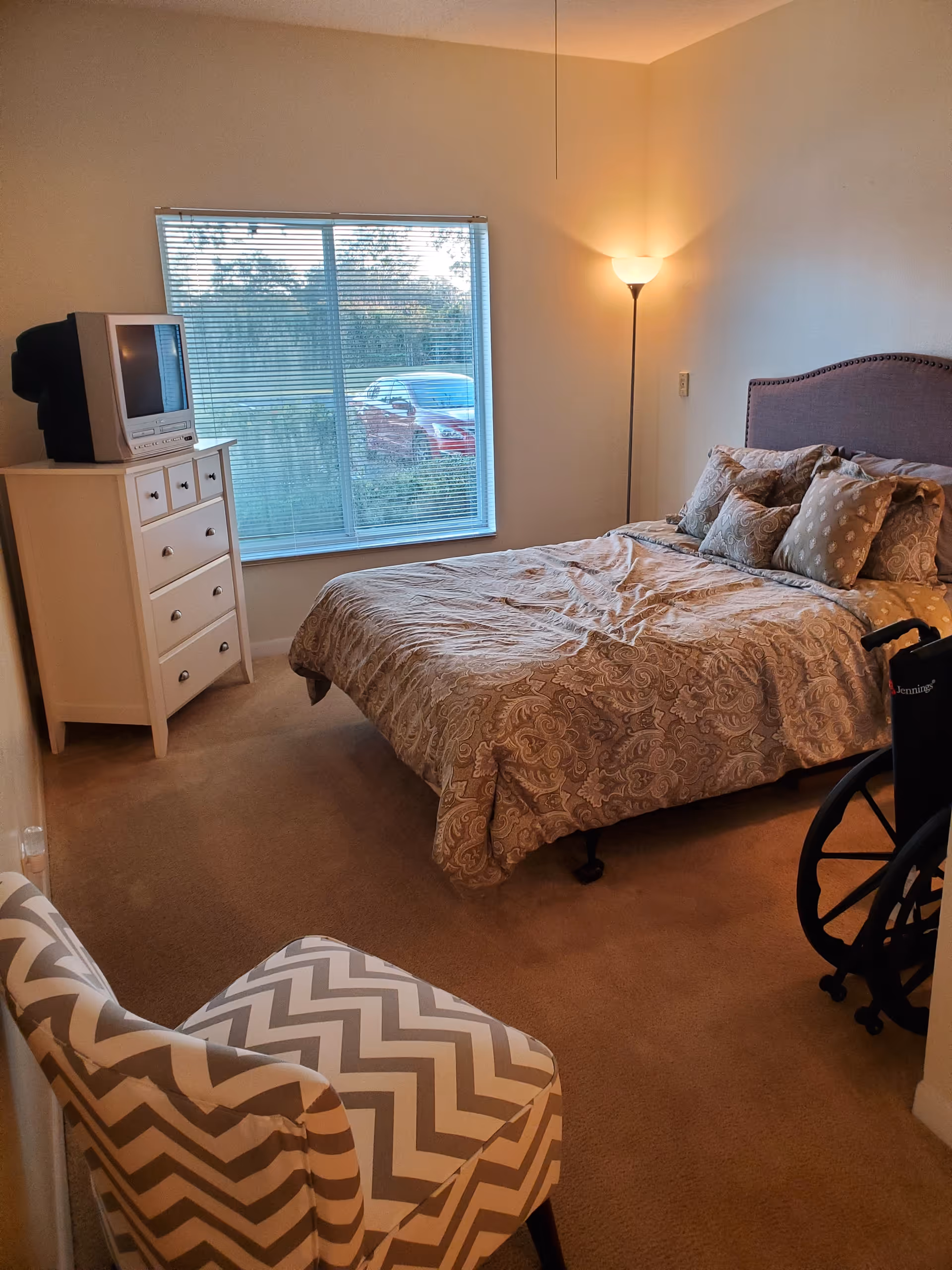 A bedroom with a bed covered in a patterned beige comforter and multiple pillows. There is a white dresser with a small TV on top near a window with blinds. A floor lamp is lit in the corner, and a wheelchair is partially visible next to the bed. A chevron-patterned chair is in the foreground.