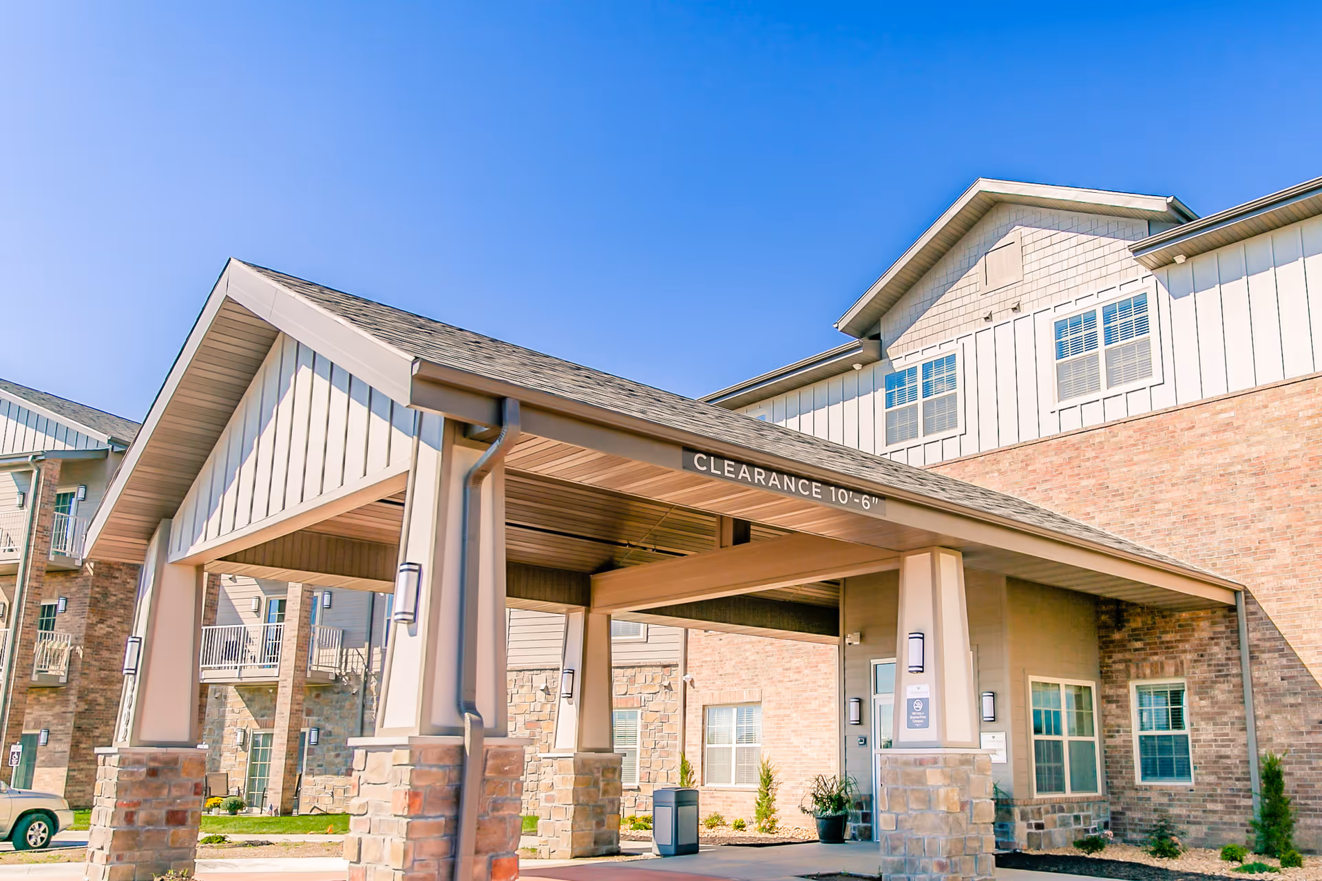 Entrance of a senior living facility with a covered drop-off area supported by stone and beige pillars, brick and siding exterior walls, and multiple windows under a clear blue sky.