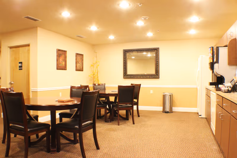 A well-lit dining area in an assisted living facility with two round wooden tables surrounded by dark wooden chairs with black cushions. The room has beige walls with a white chair rail and carpeted floor. On the right side, there is a kitchenette with a refrigerator, coffee machine, and cabinets. A large decorative mirror hangs on the wall, and two framed pictures are on the adjacent wall. A small trash can is placed near the wall under the mirror.
