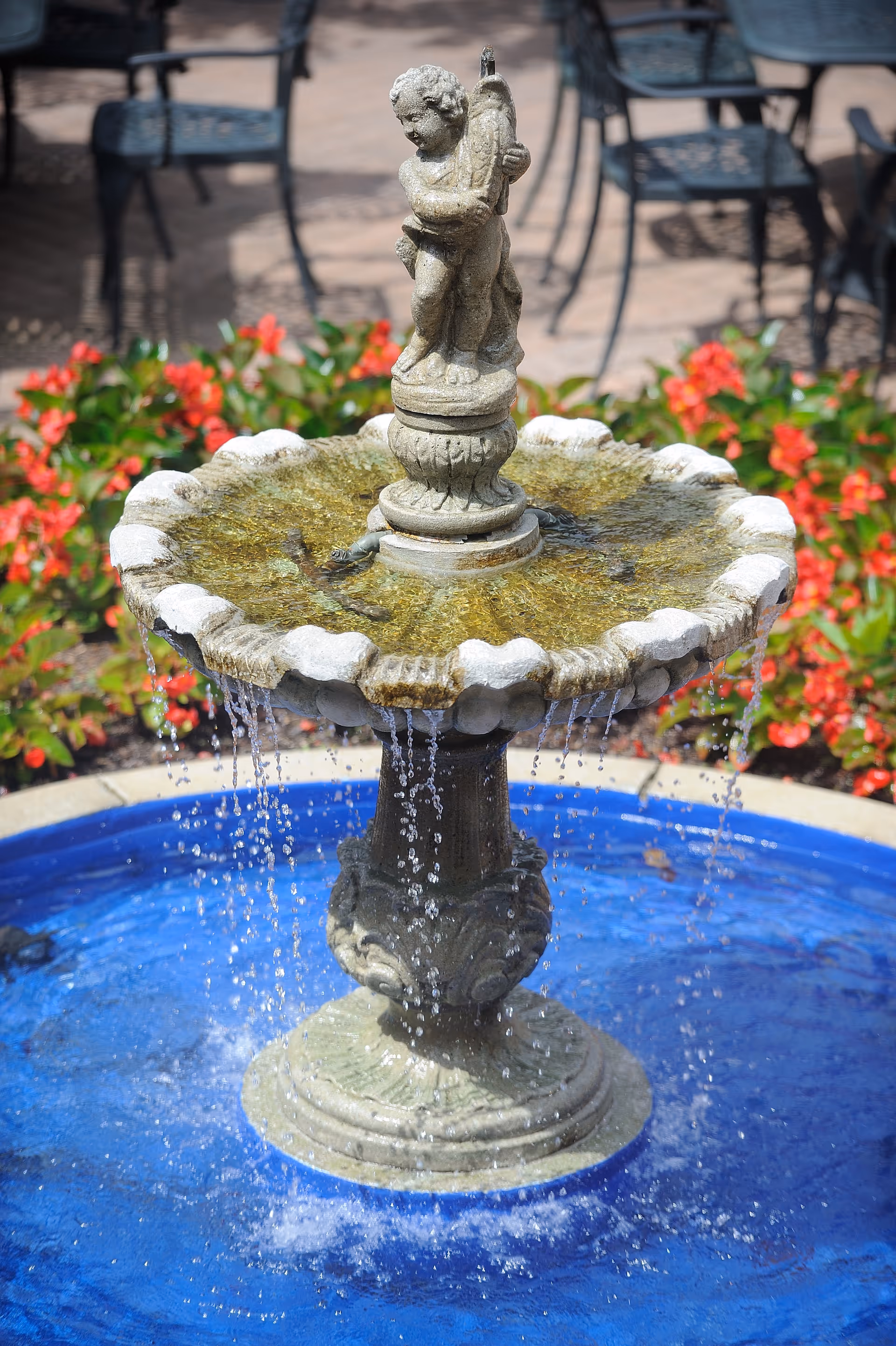 A decorative outdoor water fountain with a statue of a cherub holding a fish at the top. Water cascades from the edges of the fountain into a blue basin below. In the background, there are red flowers and black metal chairs and tables on a patio.