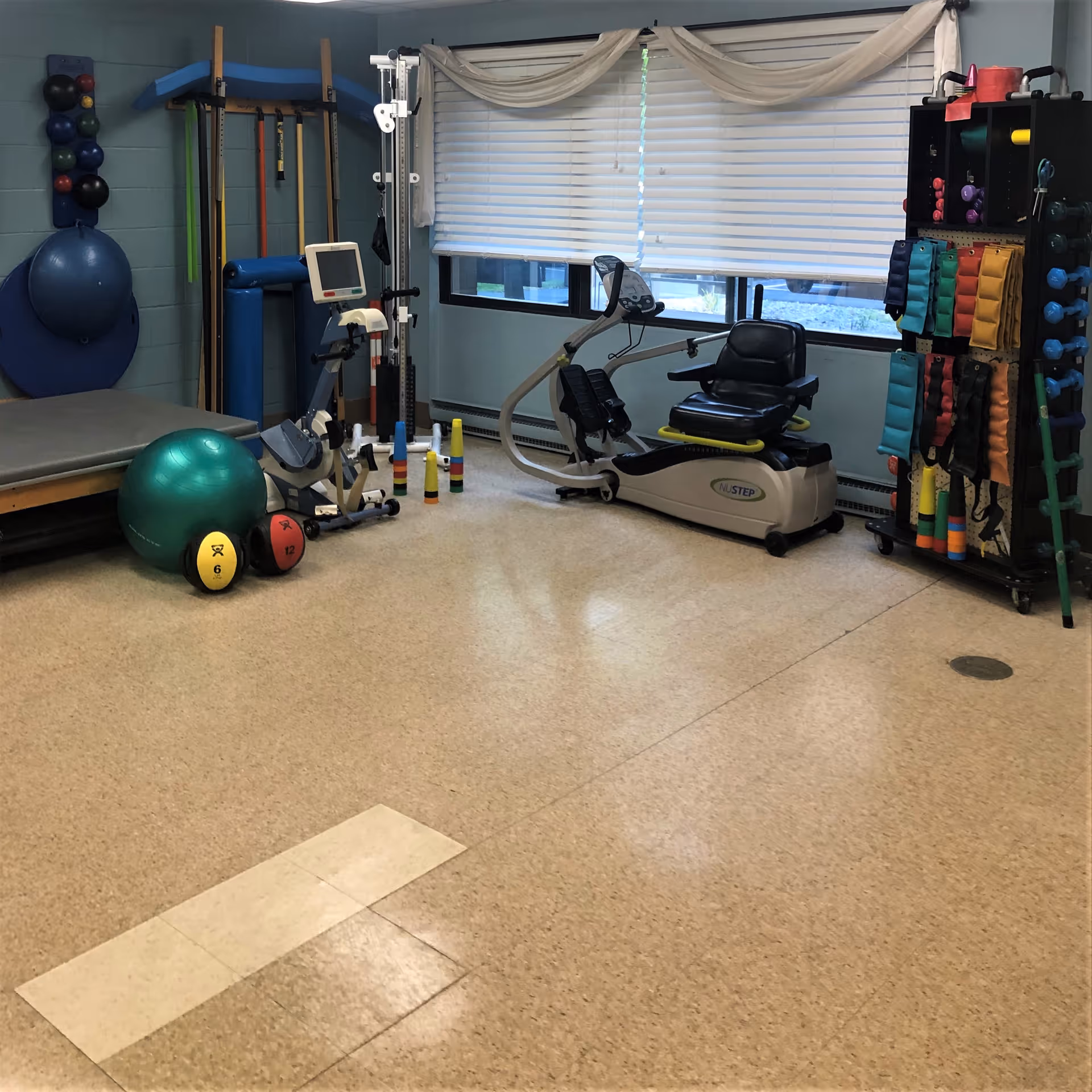 A rehabilitation room with various exercise equipment including a stationary recumbent bike, exercise balls, dumbbells, resistance bands, and other physical therapy tools. The room has light blue walls, a large window with white blinds, and a padded therapy table.