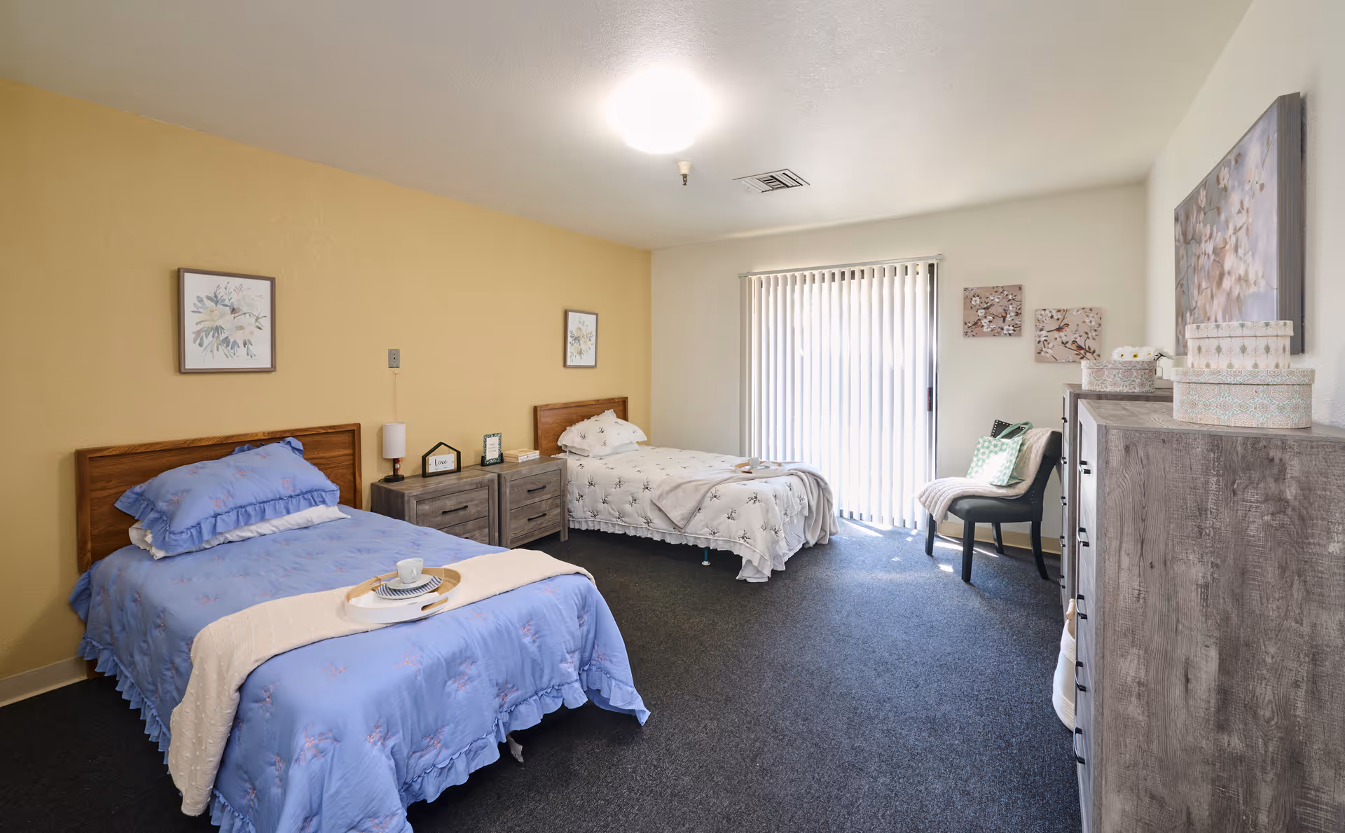 Well-lit shared bedroom with two twin beds, a nightstand, dresser, chair, and sliding vertical-blind door.