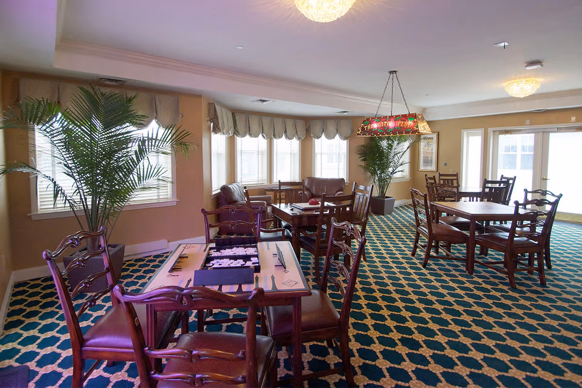A well-lit common area with multiple wooden tables and chairs arranged on a patterned carpet. There are two large potted plants near the windows and a stained glass hanging light fixture above one of the tables. The room has large windows with beige valances and a set of glass doors letting in natural light.