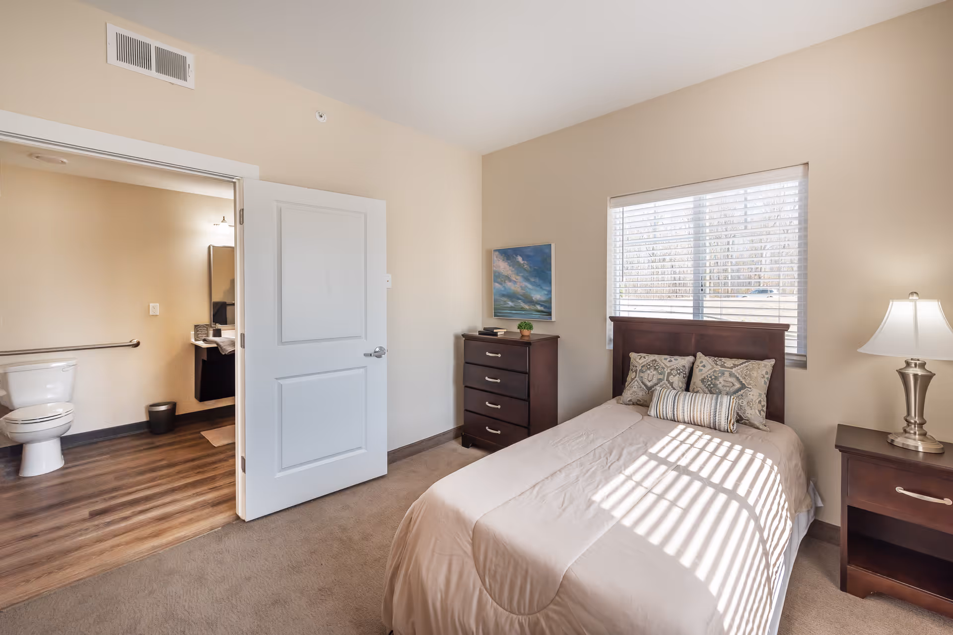 Sunlit furnished bedroom with a single bed, nightstand and dresser, and an open door showing an accessible bathroom.