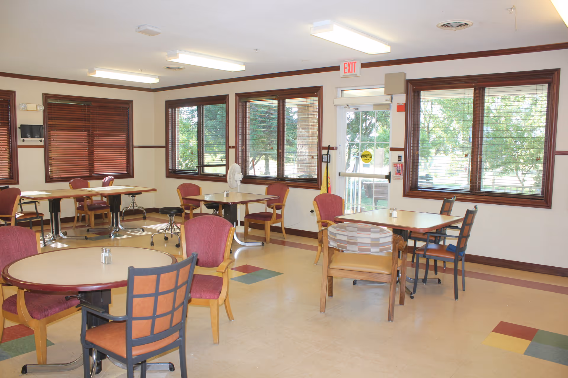 A bright dining room with multiple tables and chairs arranged around the room. The room has large windows with wooden blinds, allowing natural light to enter. The floor has a pattern with colored tiles, and there is an exit door with a glass panel. The walls are light-colored with wooden trim.