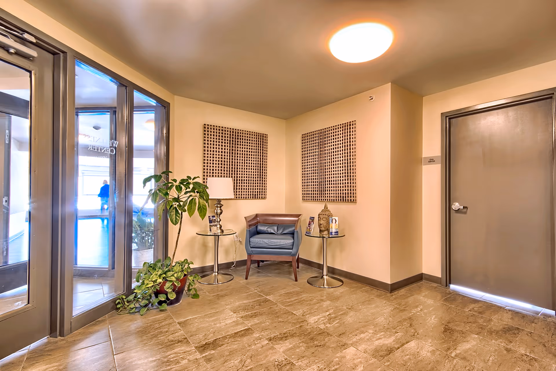 Lobby seating area with a blue armchair, two side tables, potted plants, and glass entrance doors.