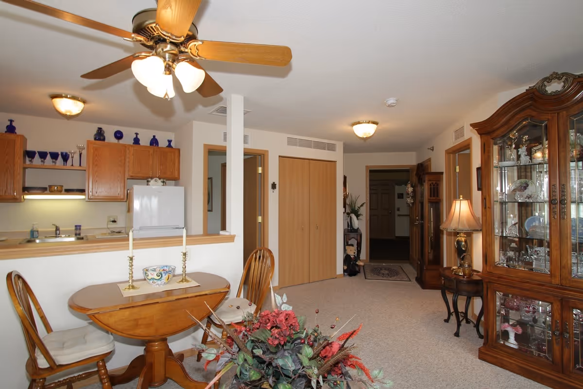 Interior view of a senior living facility showing a small dining area with a wooden table and two chairs, a kitchen with wooden cabinets and a white refrigerator, a ceiling fan with lights, and a living area with a glass display cabinet filled with decorative items. There is a hallway with closed wooden doors and a lamp on a side table.