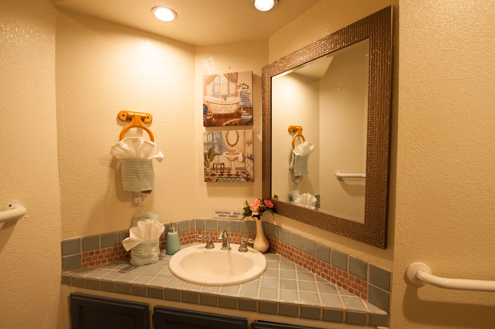A small bathroom sink area with a tiled countertop and backsplash. Above the sink is a large rectangular mirror with a textured frame. On the countertop, there is a soap dispenser, a small vase with pink flowers, and a tissue holder with tissues. A towel ring with a folded towel and tissues is mounted on the wall to the left of the mirror. Two framed pictures depicting vintage bathroom scenes hang on the wall above the sink. The walls are painted beige, and there is a grab bar visible on the right side of the image.