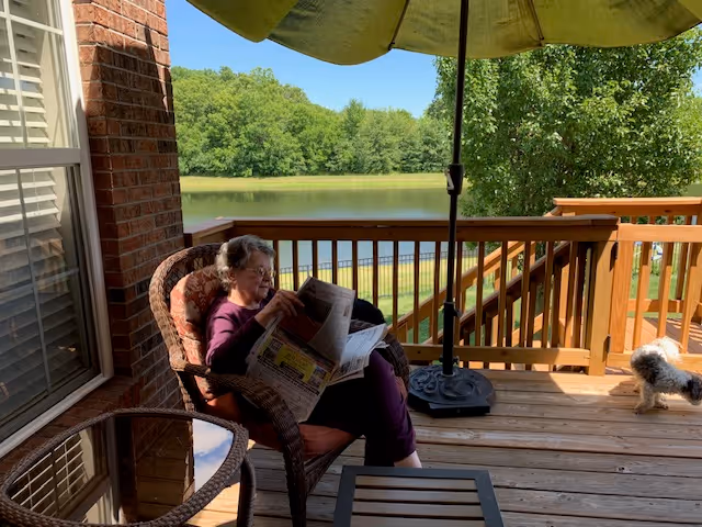 An elderly woman sitting on a cushioned wicker chair on a wooden deck, reading a newspaper. The deck overlooks a serene pond surrounded by green trees. There is a large green patio umbrella providing shade, a small round glass table, and a small dog walking on the deck.