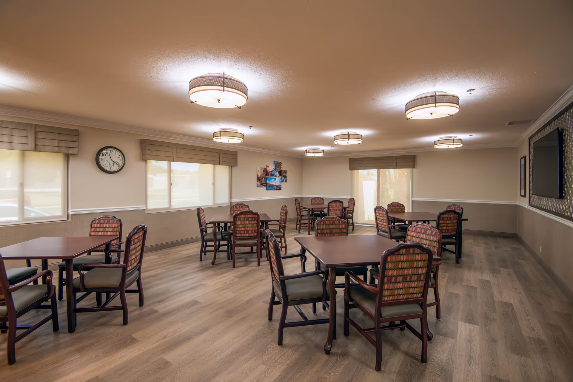 Spacious senior dining/activity room with multiple square tables and upholstered chairs, large windows, a wall clock, and ceiling lights.