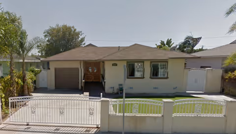 Front exterior view of a single-story residential building with a gated driveway, a garage on the left, and two windows on the right side. The building is surrounded by a low wall with a metal gate and some greenery including palm trees and bushes.