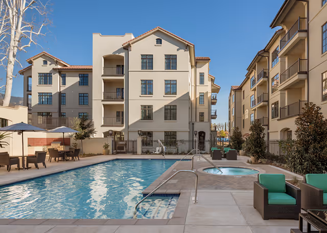 Outdoor courtyard featuring a swimming pool, hot tub, lounge seating and multi-story apartment buildings under a clear blue sky.