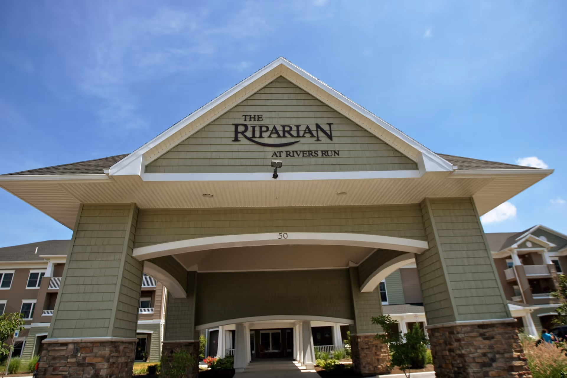 Entrance of a senior living facility named The Riparian at Rivers Run, featuring a large covered driveway with green siding and stone pillars under a clear blue sky.