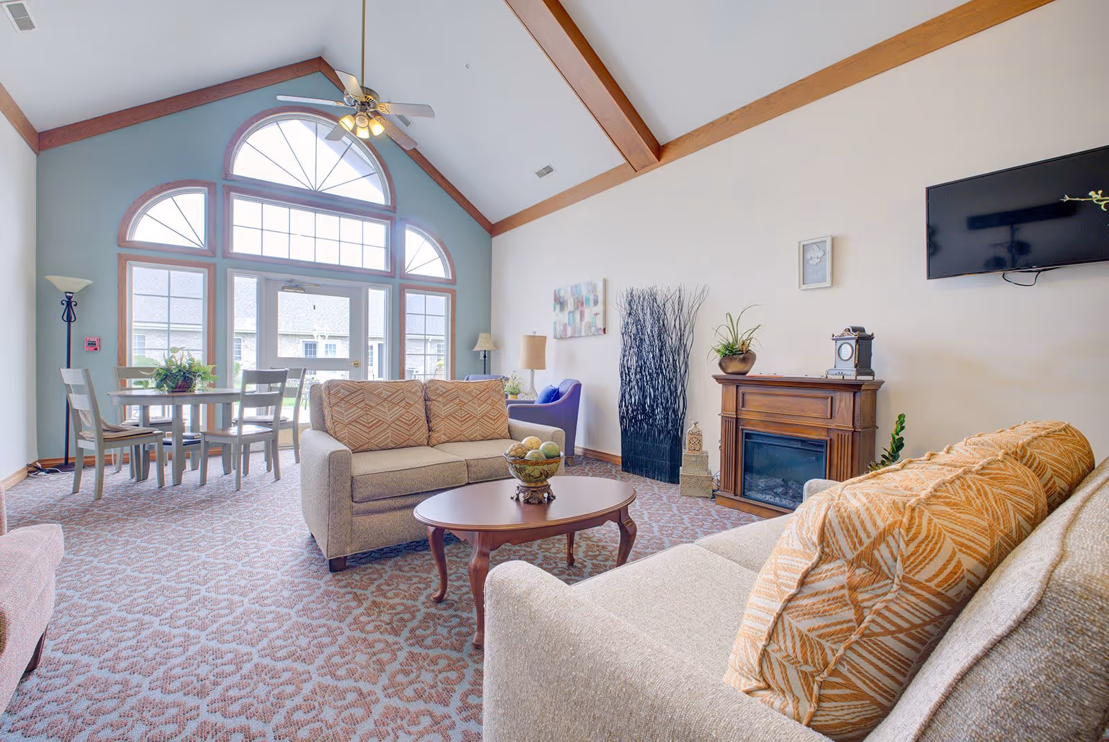 A bright and spacious living room with high vaulted ceilings and large arched windows letting in natural light. The room features two beige sofas with patterned cushions, a wooden coffee table with a decorative bowl, a small dining table with chairs near the windows, a wall-mounted TV, and a wooden electric fireplace. The carpet has a subtle floral pattern, and there are decorative plants and artwork on the walls.