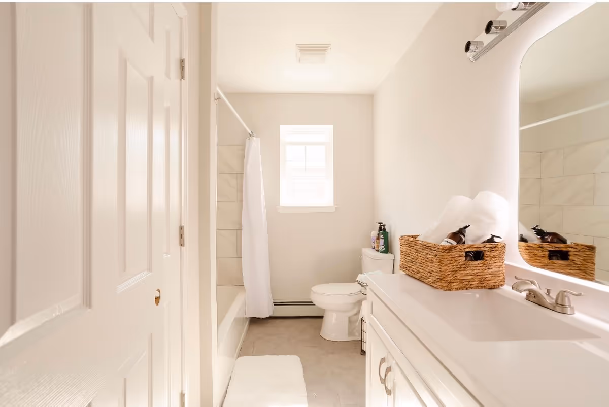 A bright and clean bathroom with a white door partially open, a bathtub with a white shower curtain, a small window above the toilet, and a white vanity with a large mirror. On the vanity, there is a woven basket holding white towels and some bottles of toiletries.