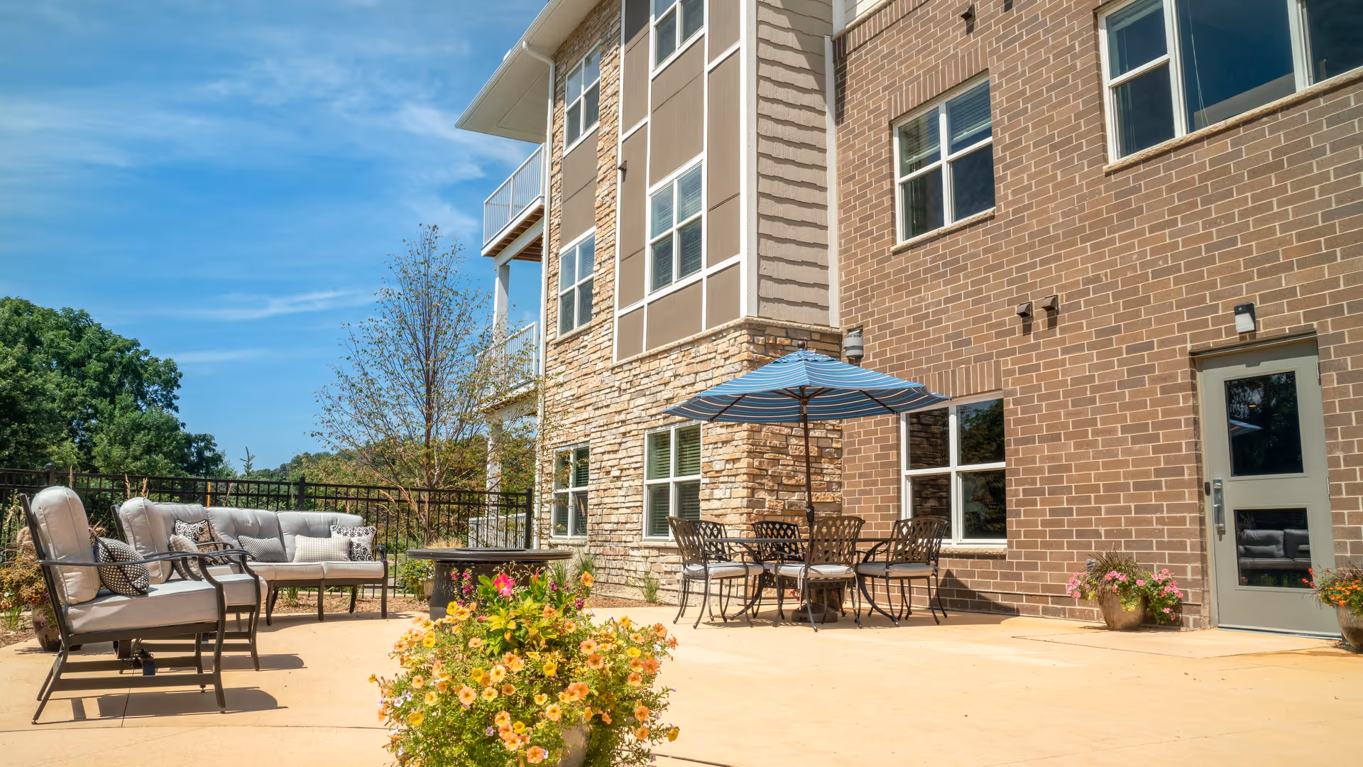 Sunlit outdoor patio with cushioned seating, a dining table under a blue umbrella, potted flowers, and the brick-and-stone exterior of the senior living building.
