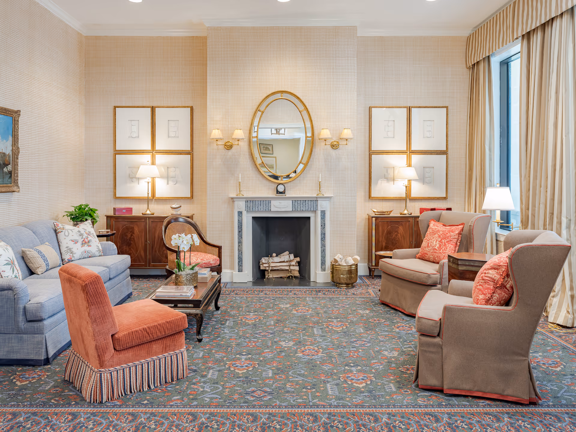 A cozy living room with a patterned carpet, a blue sofa with floral pillows, an orange upholstered chair, two beige armchairs with red cushions, a wooden coffee table with a flower vase, and a fireplace with a gold-framed oval mirror above it. The walls have light-colored wallpaper and framed artwork, and there are two wooden cabinets with table lamps on either side of the fireplace. A large window with beige curtains lets in natural light.