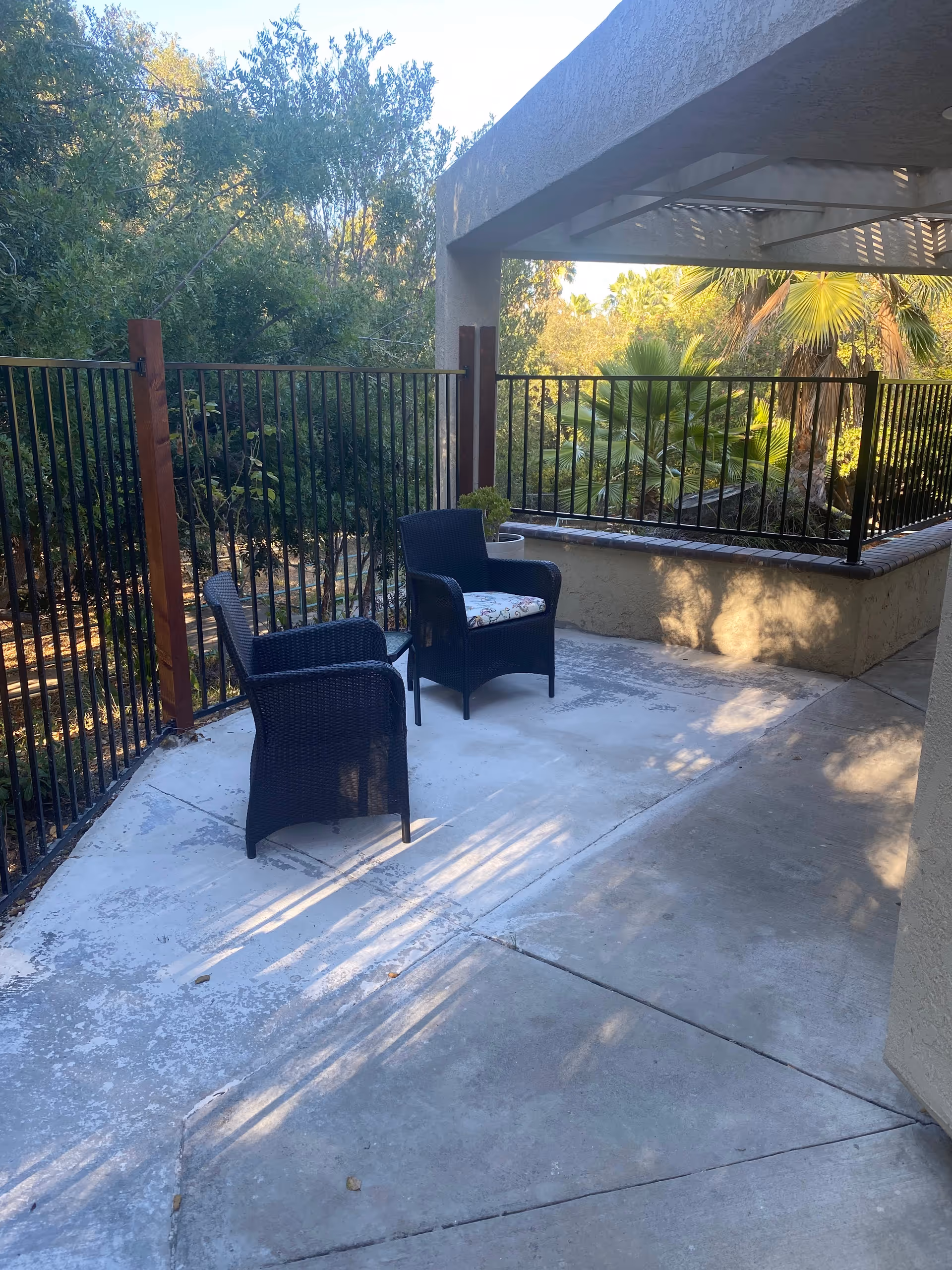 Outdoor patio area with two black wicker chairs, one with a floral cushion, surrounded by a black metal fence and greenery including trees and palm plants in the background.