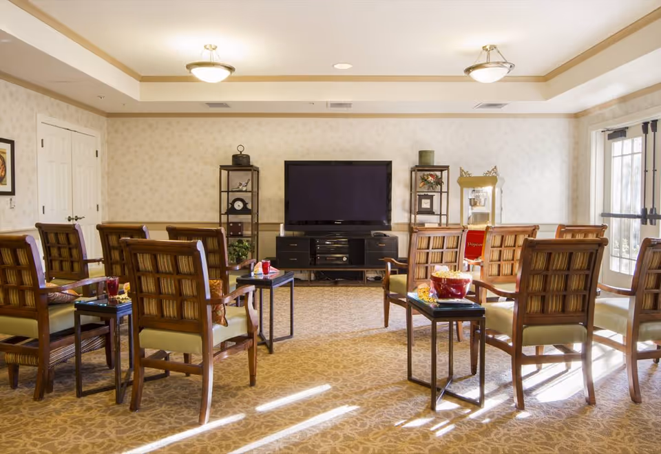 A bright and spacious common living area with multiple wooden chairs arranged in two rows facing a large flat-screen TV mounted on a black entertainment unit. There are small side tables with snacks and drinks, a popcorn machine in the corner, and decorative shelves with plants and ornaments. The room has patterned carpet, light-colored wallpaper, and ceiling lights.